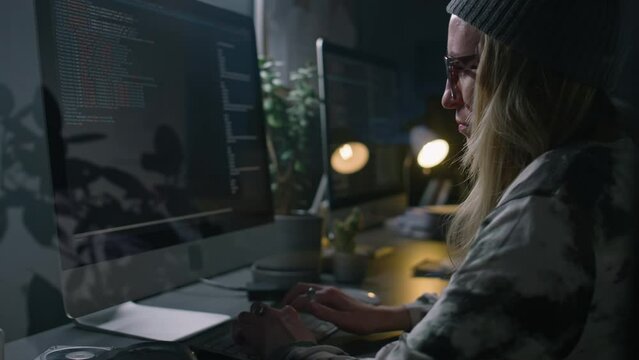 Medium close-up view through glass wall of Caucasian female software developer in beanie, glasses sitting at desk in dark company office at night, typing on keyboard, writing computer code for project