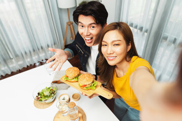 Top view of smiling host chef influencers taking photo with smartphone by selfie holding double hamburger on cooking show special easy with salad side dish homemade on social media channel. Infobahn.