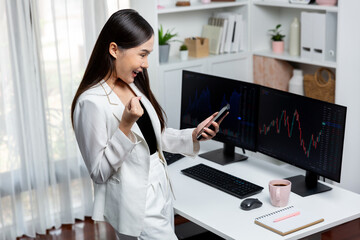 Successful young Asian businesswoman earning high profit raising fist up, looking smartphone for exchange stock market's financial technology with dynamic business graph at modern office. Stratagem.