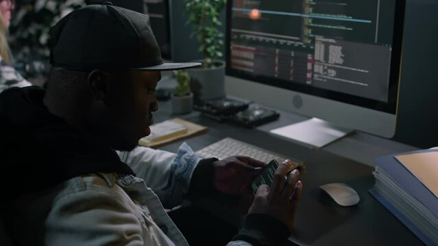 Medium close-up of young African American male mobile application developer in hoodie and baseball cap sitting at desk in office, writing computer code, then checking for errors on smartphone