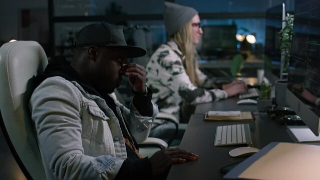 Medium side of tired young African American male software developer in hoodie, baseball cap writing computer code in dark startup office at night, getting errors, rubbing sore eyes and forehead