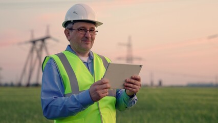 Power engineer in safety helmet checks power line, digital tablet. Construction engineer looking at camera smiling, working with tablet outdoors, specializing in electricity supply works outdoors.