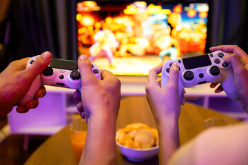Focus on hands couple or friends joyful player video game on TV using joysticks together with blurry screen. Fronted snack and drinks on table in studio room in neon light at comfy home. Postulate.