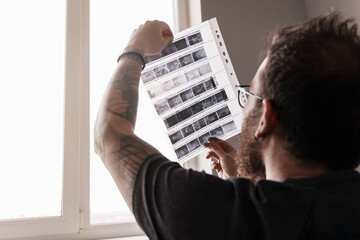 Close-up of a bearded photographer holding and inspecting film negatives in natural light by a window.