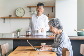家でパソコンを使う高齢者女性とキッチンにいる高齢者男性

