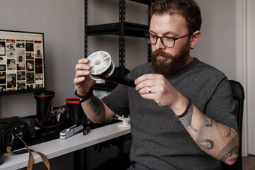 A focused male photographer reviews film negatives in his workspace filled with photography equipment and a computer.