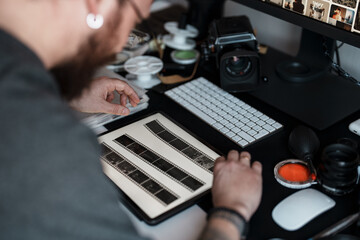 Focused photographer examines film strips amidst a modern workspace setup with a digital workstation and vintage camera equipment.
