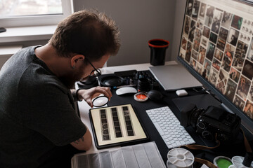 Focused male photographer analyzing film negatives with a magnifying glass at his modern workspace.