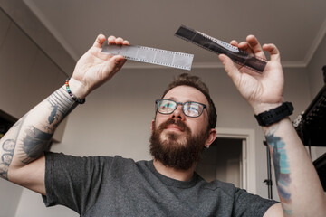 A bearded man with tattoos holds up and examines two strips of film negatives, comparing their details in a well-lit room.