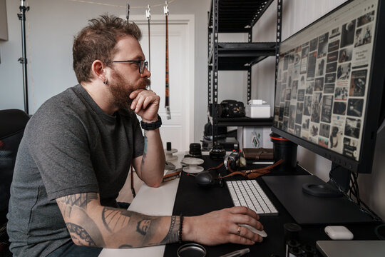 Focused male photographer with tattoos analyzing photographs on a widescreen monitor in a well-equipped home studio.
