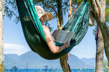 View of a woman in a hammock reading a book in front of a lake and big mountains