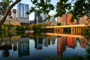 Golden hour in Austin, Texas. The cityscape reflected on the surface of Lady Bird Lake, accompanied by the gliding of a river boat tour. Perspective from the waterfront's edge during a summer evening.