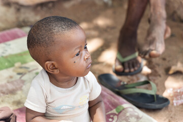 village african baby with a fly on the cheek sitting on the ground on a blanket in the yard
