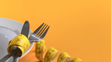 Closeup measuring tape around fork and knife on yellow background. Weight loss and healthy concept