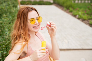 Young red-haired woman blowing soap bubbles outdoors. Girl with braces on her teeth. 