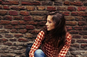 Beautiful brunette woman with long hair sitting near the old brick wall building background in casual red shirt on the spring city. Closeup vintage portrait of model. Profile