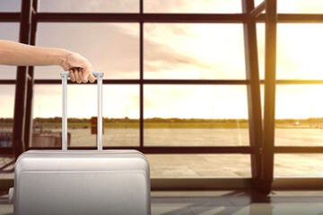 Image of man hand holding soft grey suitcase with airport view on background.