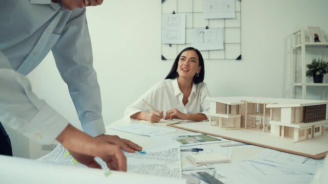 Skilled caucasian interior designer working with house model while discussing with asian architect engineer about house plan on meeting table with blueprint and house model scatter around. Immaculate.