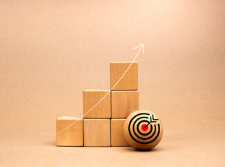 Target icon on wood ball near wooden cube blocks bar graph chart steps with rise up arrows on beige...