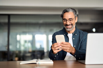 Smiling older Indian business man sitting at desk using mobile phone. Happy mature businessman executive, busy middle aged entrepreneur investor looking at smartphone working in office with cellphone.