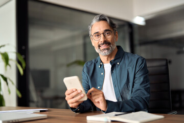 Middle aged businessman looking at camera working holding smartphone. Mature 45 years old business man executive, business owner or entrepreneur sitting at desk using mobile cell phone. Portrait.