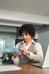 Young busy professional young African American business woman employee holding cellphone using mobile looking at cell phone technology sitting at desk in office working on laptop, Vertical photo.