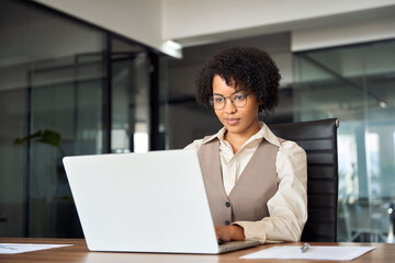 Busy African American business woman using laptop working in corporate office. Smiling young businesswoman executive at work, professional female company hr or bank manager looking at computer.