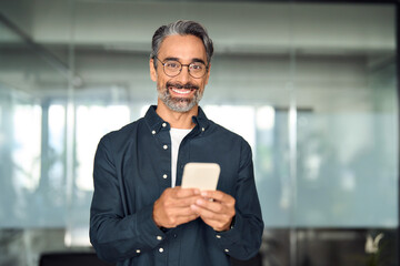 Happy mature business man executive using smartphone standing in office. Portrait. Middle aged businessman entrepreneur leader holding mobile cell phone looking at camera at work.