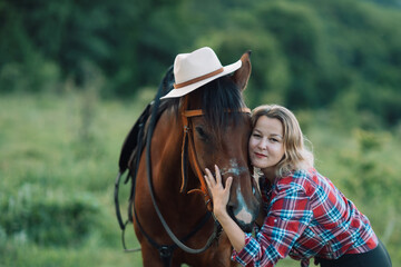 Happy blonde with horse in forest. Woman and a horse walking through the field during the day. Dressed in a plaid shirt and black leggings.