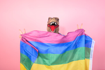 non-binary person with dinosaur head waving a rainbow flag on an isolated pink background