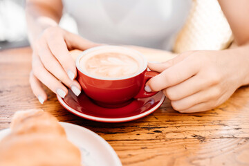 A woman is holding a red coffee cup with a white saucer. The woman is sitting at a table with a plate of croissants in front of her.