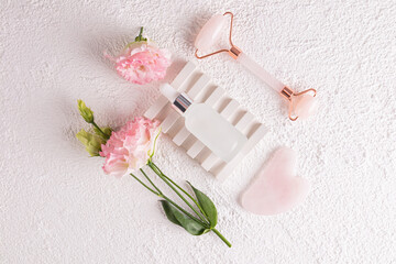 A white matte bottle of cosmetics, a rose quartz massage roller and a gua sha scraper lie on an embossed podium with pink flowers Top view