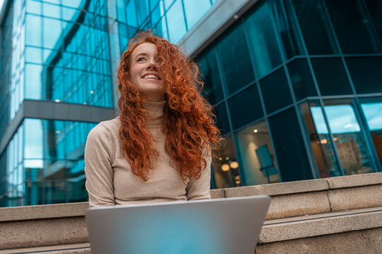 Portrait of beautiful smiling lady reading e-mails on  laptop while sitting on steps outside of the business centre