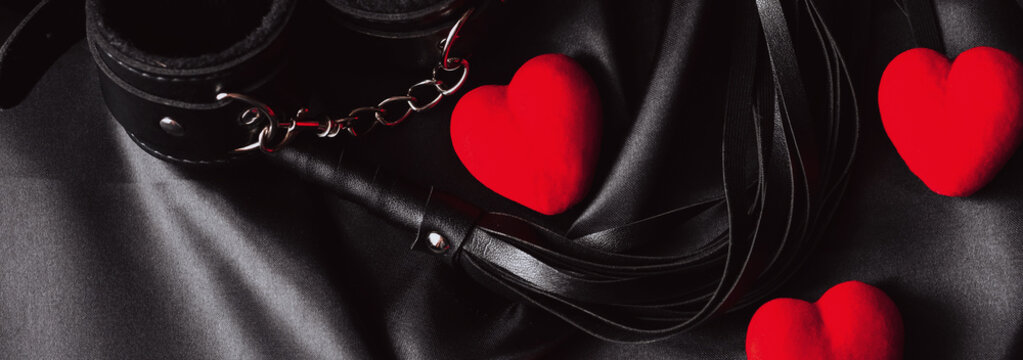 set of BDSM sex toys with handcuffs, whip flogger with red hearts for submission and domination on a black background. Gift for Valentine's day. Wide header cover for banner for sex shop
