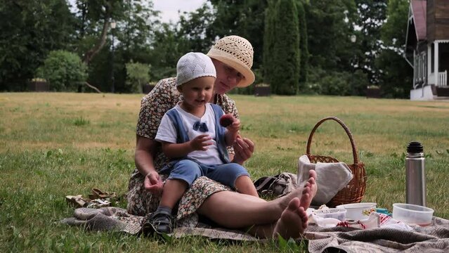 A young woman and her two-year-old child were picnicking on green grass in park. Mother and son eating a peach during a family meal. Barefoot girl holding a child in her arms and helping him to eat.