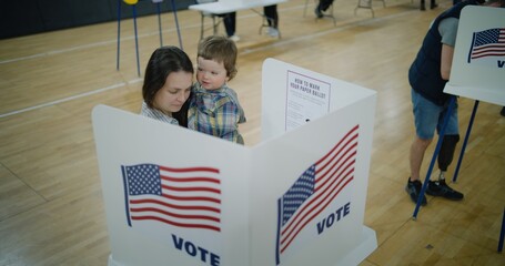 Female voter with baby on hands stands at voting booth. American citizens come to vote in polling...