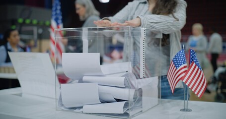 Close up shot of polling box standing on table in polling station. American female voter with kid...