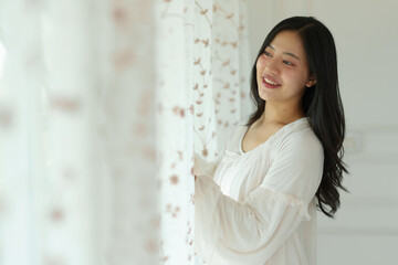 Fototapeta na wymiar Asian woman in white pajamas is opening the curtains and looking out the window smiling in her room in the morning. Soft sunlight. Vacation, lifestyle concept.