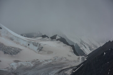 Dramatic alpine view to large glacier and sharp rocky peaked top in gray low clouds in harsh...