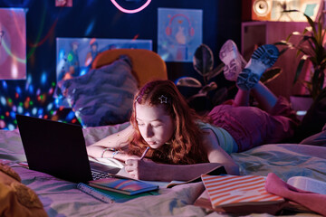 Modern teen girl lying on bed in her bedroom and doing homework task, writing in notebook late in...
