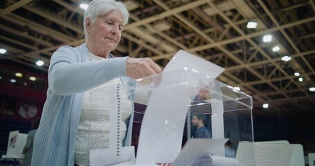Close up shot of polling box standing on table in polling station. Elderly female voter puts bulletin into box, looks at camera. Political races of US presidential candidates. National Election Day. - obrazy, fototapety, plakaty