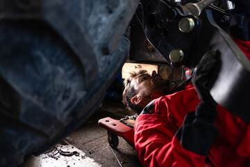 Experienced caucasian mechanic working under the tractor repairing agricultural machinery.