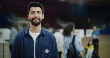 Portrait of Caucasian man with badge smiling and looking at camera standing at polling station....