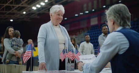 Multi ethnic American citizens come to vote in polling station. Elderly woman talks with polling officer and takes vote bulletin. Political races of US presidential candidates. National Election Day. - obrazy, fototapety, plakaty