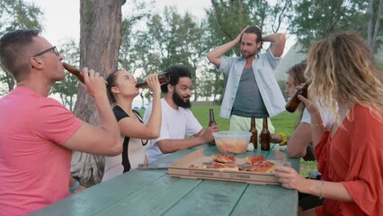 Lively group of friends toasting with beer bottles, sharing a moment of celebration at a sunlit outdoor table, epitomizing the joy of casual gatherings. Slow motion. 