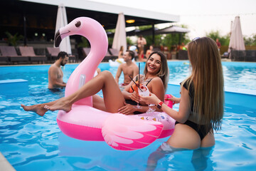 Pretty woman in bikini swimwear hanging out on inflatable pink flamingo mattress on night pool party. Friends partying with cocktails in resort villa swimming pool. Girls toast, cheer with drinks.