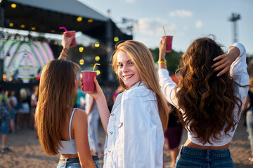 Group of joyful women cheers with drinks, enjoying live music at sunset beach festival. Smiling...