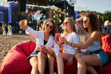 Group of women enjoy music fest on beach, lounging on bean bags. Casual attire, sunglasses, sipping...
