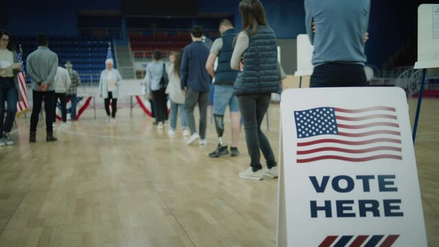 Queue of multi ethnic American citizens come to vote in polling station. Vote here sign on the floor. National Election Day in United States. Political races of US presidential candidates. Dolly shot.