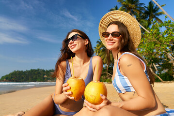 Two women in swimsuits and sunglasses sit on tropical beach holding fresh coconuts. Enjoying sun,...